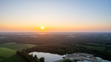 Bir hava manzarası, kırsal bir araziyi süsleyen sakin bir gün doğumunu yakalar, geniş alanlar, sakin bir göl ve uzak bir ağaçlık alana sıcak bir ışıltı yayar. Ufuk yumuşak pastel renklerle süslenmiş.