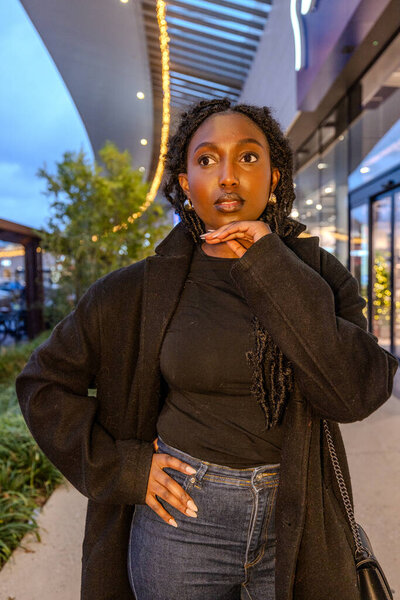 This image captures a young Black woman in a moment of contemplation, her gaze directed upwards as if pondering a profound thought or decision. The urban evening serves as a luminous backdrop, with a