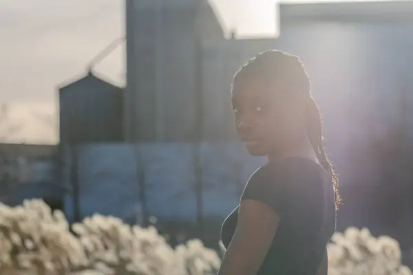 This image captures a young Black woman glancing over her shoulder, her face gently illuminated by the soft glow of a setting sun. Behind her, the silhouettes of city buildings rise against the sky