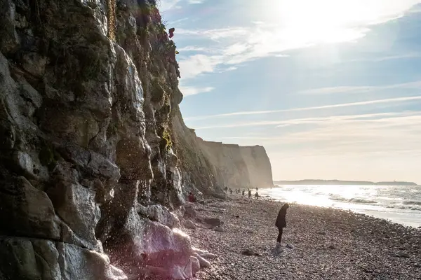 A striking image that tells a story of exploration and adventure along a rugged cliffside. The focus here is on the figure walking parallel to the cliffs face, which is bathed in sunlight