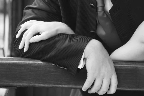 This image, rendered in black and white, captures a quiet, yet profound moment as two hands lay intertwined, resting on a wooden bench. The male hand, dressed in a pinstripe suit, conveys a sense of