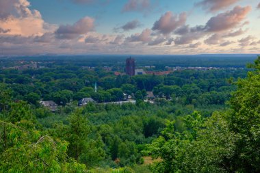 Bu görüntü, dinamik bir şafak vakti altında yemyeşil ortamda kurulmuş banliyö yayılımının nefes kesici panoramik bir görüntüsünü sunuyor. Odak noktası, ağaç tepelerinin üzerinde yükselen önemli bir bina.