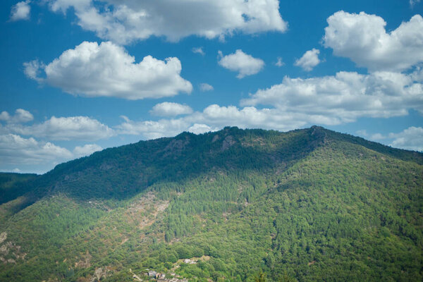 This landscape photograph features the lush, green mountains of the Ardeche region under a sky dotted with fluffy cumulus clouds. The play of light and shadow across the mountainside creates depth and