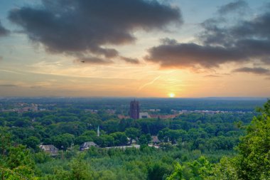 Bu sakin görüntü günbatımında banliyö manzarasının geniş bir görüntüsünü sunuyor. Durgun güneş ufka doğru batar, yeşil ağaçların tepelerini süsleyen yumuşak, ılık bir parıltı saçar ve