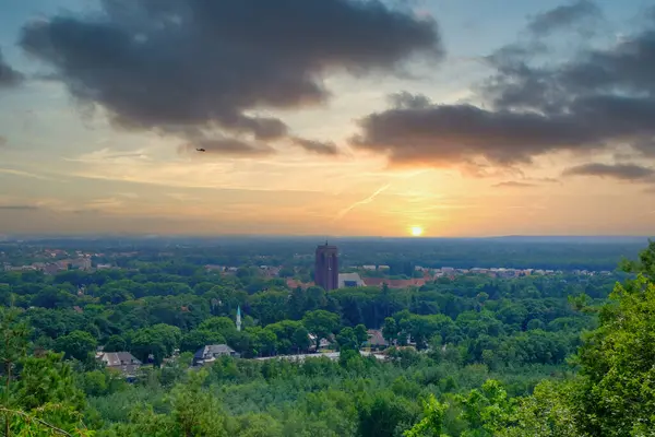 Bu sakin görüntü günbatımında banliyö manzarasının geniş bir görüntüsünü sunuyor. Durgun güneş ufka doğru batar, yeşil ağaçların tepelerini süsleyen yumuşak, ılık bir parıltı saçar ve