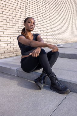 Görüntüde, beton basamaklarda oturan genç bir Afrikalı kadın tuğla bir arka planda görülüyor. Rahat ve dalgın görünüyor, sakin ve kendinden emin bir şekilde doğrudan kameraya bakıyor.