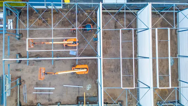 Bu hava görüntüsü bir inşaat alanının kuş bakışı görüntüsünü sağlıyor, yapı iskelesi ve makinelerin geometrik düzenini gösteriyor. İki turuncu teleskopik patlama asansörü çamurlu zemine karşı duruyor.