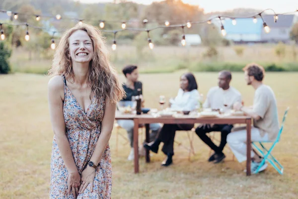 This image features a young woman in the foreground, standing in a grassy outdoor area adorned with string lights above. She is wearing a floral dress and is smiling broadly, showcasing a look of