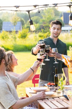 Bu görüntü, arkadaşlarınızın kırsal ahşap bir masanın etrafında toplanıp, açık havada kırmızı şarap kadehleriyle kadeh kaldırırken ki şenlik ruhunu özetliyor. Batan güneş sıcak ve davetkar bir hava yaratır.