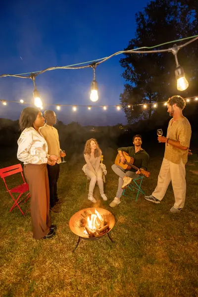 The image portrays a group of friends gathered in a backyard around a fire pit under the ambient glow of string lights. The focus is on social interaction and the enjoyment of an acoustic guitar