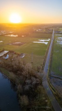 Fotoğraf günbatımında kırsal bir manzaranın havadan görüntüsünü sunuyor. Çerçevenin içinden geçen, elektrik direkleriyle çevrili düz bir yol, insan yerleşimini ve doğanın ortasındaki altyapıyı gösterir.