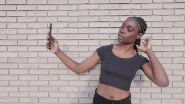 このストック映像は 編組された髪のアフリカ系アメリカ人女性がトップとジーンズを着て 白いレンガの壁の背景と彼女のスマートフォンと対話しています ファーストシーン — ストック動画