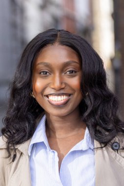 Fotoğrafta parlak ve çekici bir gülümsemesi olan genç bir Afrikalı Amerikalı kadın görülüyor. Açık havada duruyor. Arka planda bulanık bir şehir manzarası var. Şehir ortamını işaret ediyor. Onun.