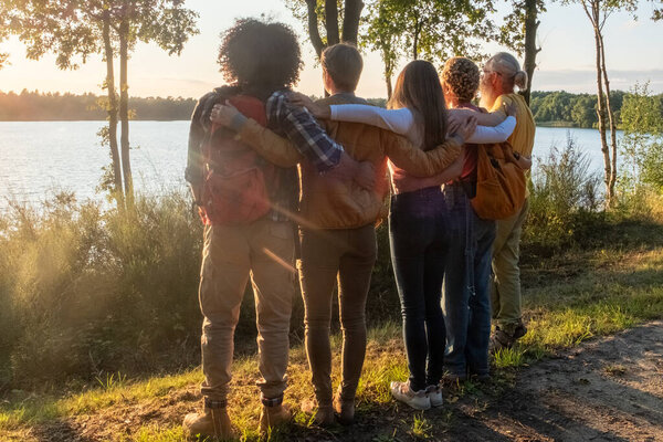 This image captures a tender and peaceful moment as a group of friends, with their backs to the camera, embrace while looking out over a tranquil lake at sunset. The setting sun casts a warm glow over