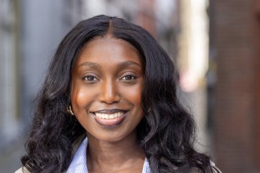 Bu yakın plan portrede parlak, çekici bir gülümsemesi olan, şehir ortamında duran genç bir Afrikalı kadın görülüyor. Doğal güzelliği asgari makyajla vurgulanır, pürüzsüz cildini ön plana çıkarır.