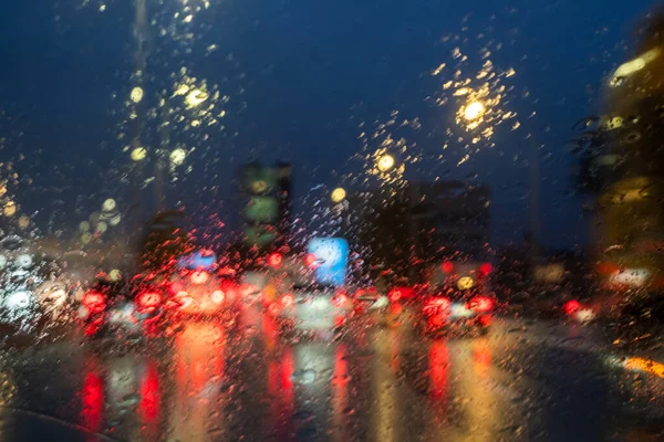 Bu görüntü, gece vakti yağmurun sıçradığı bir ön camdan görülen şehir trafik manzarasının bulanık ışıklarını yakalıyor. Camdaki damlacıklar kırmızı fren lambaları üzerinde yumuşak bir odak etkisi yaratıyor.