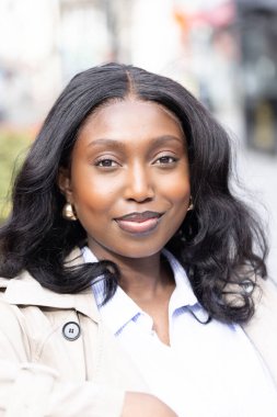 Fotoğrafta genç bir Afrikalı kadının davetkar ve sıcak bir gülümsemesi var. Açık havada, arkasında şehir manzarası bulanık. Bu da canlı bir şehir manzarası olduğunu gösteriyor. Koyu, dalgalı