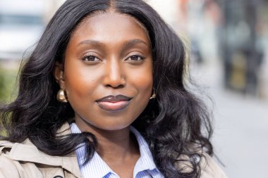 Bu yakın plan, genç bir Afrikalı kadını şehir ortamında güven ve zarafet yayarken yakalıyor. Klasik bir trençkot ve mavi çizgili bir gömlek giyiyor.