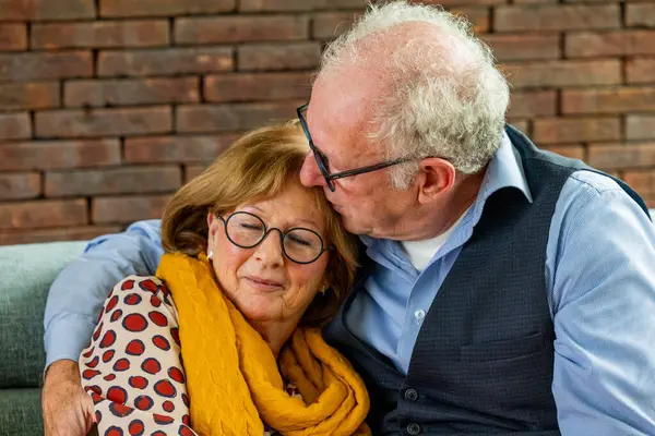 stock image In this intimate scene, an elderly man with grey hair and glasses is depicted giving a tender kiss on the forehead to his partner. The woman, wearing stylish glasses and a scarf, closes her eyes and