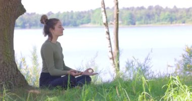 Bu sakin, yavaş çekim görüntüsü, dingin bir göl kenarında derin meditasyon yapan genç bir kadını yakalıyor. Bir ağacın gölgesinin altında konumlanmış ve manzaranın doğal güzelliğiyle çevrelenmiş.