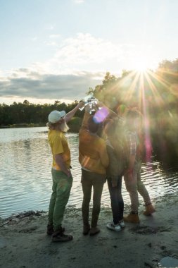 Bu görüntü, göl kenarında toplanmış bir grup arkadaşı bir kutlama jesti ile ellerini kaldırırken, muhtemelen su şişeleriyle tezahürat yaparken ya da kadeh kaldırırken yakalıyor. Güneş onların ardından batıyor, ışınlarıyla.