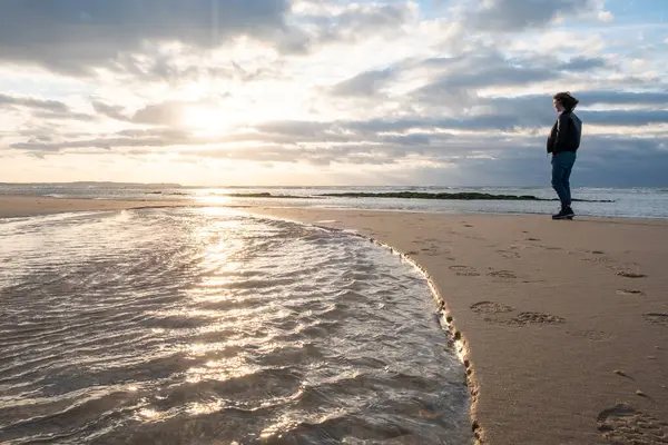 This image eloquently captures a lone individual standing on the beach, gazing out towards the sea under the soft light of the setting sun. The suns reflection on the water creates a path of light
