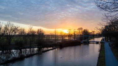 Bu sakin görüntü, sakin bir kanalda gün batımını yakalar. Güneş gökyüzüne doğru batar ve ufukta canlı turuncu bir renk bırakır. Yapraksız ağaçların silueti suları yansıtır.