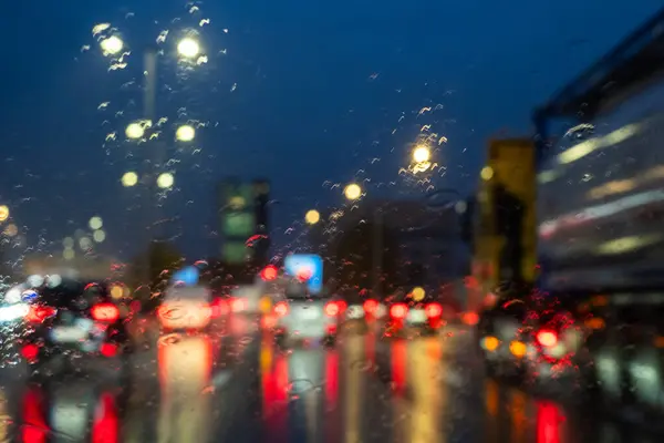 This image presents a drivers view through a rain-speckled windshield during evening rush hour. The glow of street lamps and vehicle tail lights create a bokeh effect, with the cool blue of the