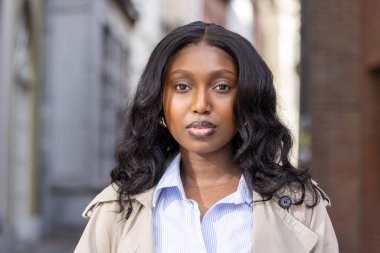 Bu görüntü, sakin ve genç bir Afrikalı kadının nazikçe kameraya baktığını gösteriyor. Mavi çizgili düğmeli bir gömlek ve açık renk bir trençkot giymesi,