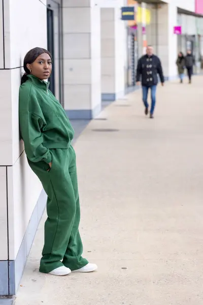The photograph depicts a young African woman with a contemplative expression, standing against a modern building on a city street. Shes wearing a coordinated green tracksuit paired with white