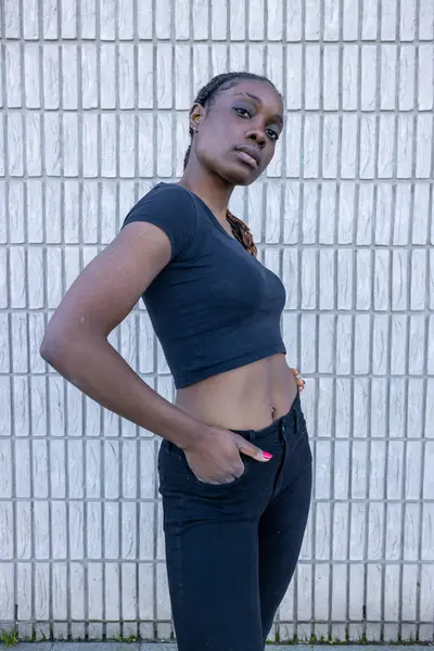 stock image This image presents a young African woman standing confidently with her hand on her hip against a white tiled wall. Her attire is a simple, fitted black crop top and jeans, complementing her assertive