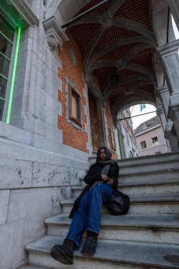 Bu görüntü, tarihi bir binanın basamaklarında dinlenmiş genç bir kadını yakalar. Kemerli geçit ve tuğla işi de dahil olmak üzere mimari ayrıntılar klasik bir Avrupa havası sağlıyor.