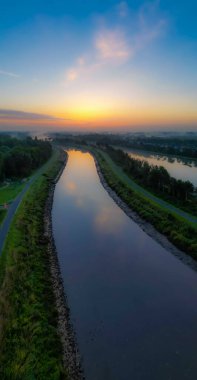Bu görüntü, sabahın erken saatlerinde bir kanalın durgun sularına yansıyan sakin bir görüntüdür. Şafak vakti hafif bir ışık gökyüzünü turuncu, sarı ve mavi renklerle aydınlatır.