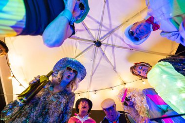 Benzersiz bir şemsiye altı perspektifi, canlı kostümlü bir yaşlılar çemberini yakalıyor, yaylı ışıklarla aydınlatılmış bir partinin tadını çıkarıyor ve şarkı söylüyor. Şemsiyenin Altındaki Dinamik Kıdemli Kostüm Görünümü