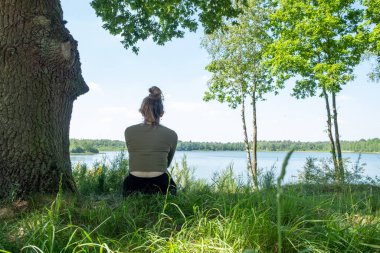 With her back to the viewer, a woman is seated on the lush grass, looking out over a still lake. The natural landscape, dotted with trees and bathed in sunlight, frames her in a moment of solitude and clipart