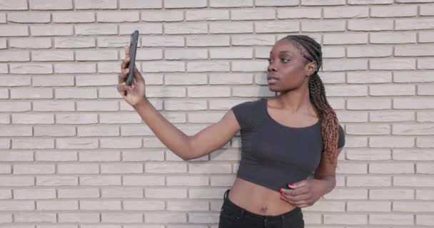 魅惑的なスローモーションシーケンスでは 若い女性がセルフィーを撮ったり スマートフォンでビデオを録画したりすることに興奮しています 彼女の焦点を当てた表現と 白いレンガのカジュアルでスタイリッシュな背景 — ストック動画