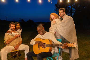 Bir grup arkadaş, gece gökyüzünün altında, battaniyelere sarılı, gitar çalarken rahat ve müzikal bir açık hava deneyimi yaratan huzurlu bir anın tadını çıkarıyorlar. Açık Hava Serenadı 'nda Rahat Gece Serenadı