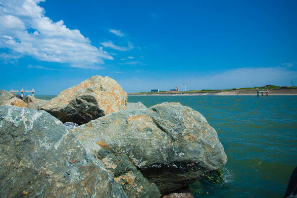Изображение представляет собой спокойную береговую линию, где большие скалы служат естественным барьером между пышной зеленой землей и безмятежной голубой водой. Пушистые белые облака танцуют над небом