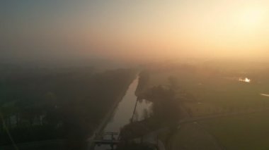Bu görüntüler bulanık bir sabahın huzur dolu özünü, dolambaçlı bir kanalda yakalıyor. Hava manzarası, güneşin sislerin arasından yavaşça yükseldiğini gösterir. Suyun kanadı ağaçlarla çevrilidir.