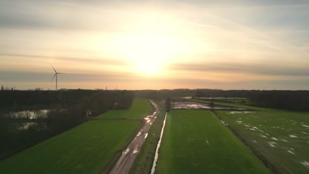 这个迷人的画面展示了日落时的和平旅程 展示了一个风力涡轮机的轮廓 旁边是一条穿过闪烁着光芒的农田的乡村道路 空中视角 — 图库视频影像
