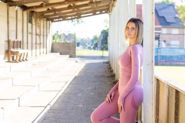A young woman clad in form-fitting pink fitness attire leans against a pillar in an urban gym setting, her gaze fixed confidently in the distance. Athletic Poise: Confident Woman in Fitness Gear at