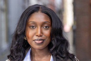 Bu görüntü özgüvenin ve özgüvenin özünü yansıtıyor. Siyah saçlı genç bir kadının yakın plan portresini yansıtıyor. Direk kameraya bakıyor, kurnaz bir gülümseme sergiliyor. Saçları.