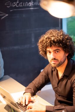 Orta Doğulu ya da İspanyol kökenli genç profesyonel bir adam, iş yerinde bir anlık konsantrasyon anında yakalanır. Kıvırcık saçlı ve siyah gömlekli, işine dalmış gibi görünüyor.