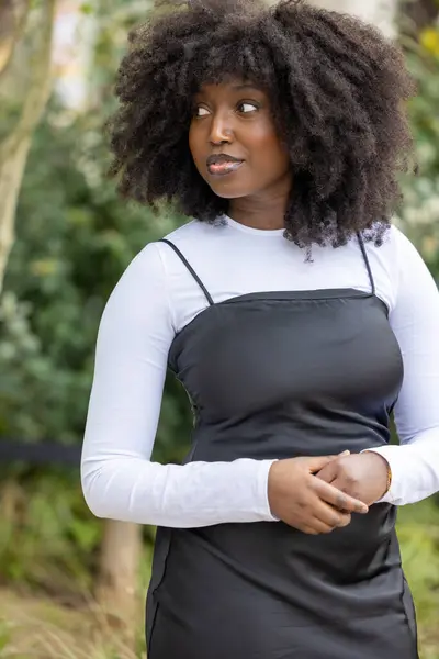 stock image A natural-light portrait of a contemplative woman dressed in a modern layered ensemble, with a black slip dress over a white long-sleeve shirt. Her expressive afro hairstyle and the soft outdoor