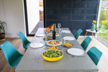 Görüntü, taze salata ve pizza da dahil olmak üzere sağlıklı gıdalarla dolu modern bir yemek masasını tasvir ediyor. Masanın temiz çizgileri ve parlak mavi sandalyeler bize bir yer veriyor.
