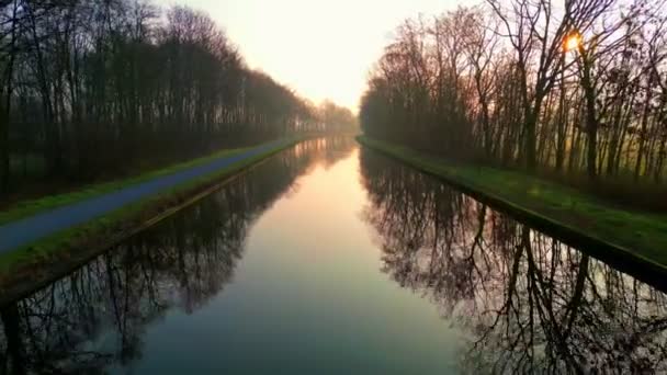 这个迷人的无人驾驶镜头捕捉了一条平静的运河的宁静之美 这条运河的两侧是一排排光秃秃的树木 反映了升起或落日的柔和光芒 平静的水面映衬着淡淡的油腻 — 图库视频影像