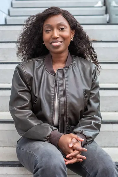 stock image A young African woman with a radiant smile sits comfortably on outdoor steps, her dark curly hair framing her face. She wears a fashionable leather jacket, presenting a relaxed yet confident posture