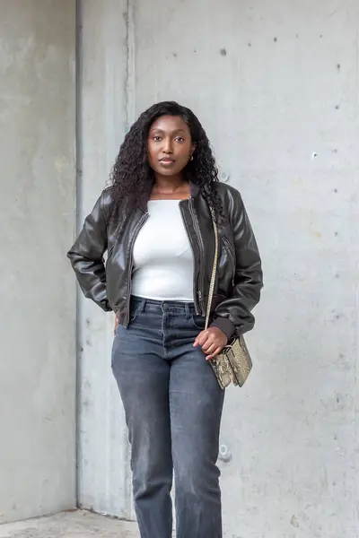 Captured against a minimalist urban backdrop, a young African woman presents a modern ensemble. Her trendy leather jacket complements the casual white top and high-waisted jeans, accessorized with a