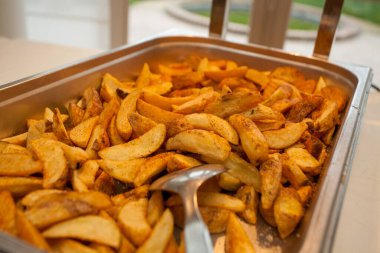 Sığ bir saha derinliği, paslanmaz çelik servis tabağındaki baharatlı patates dilimlerinin üzerinde servis yapan bir tong ile vurgulanmasını sağlar. Arka plan hafifçe bulanıklaşıyor, gayri resmi bir olay ayarını işaret ediyor