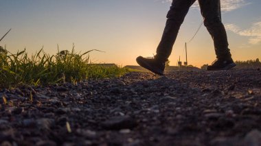 Bu görüntü, gün batımında kırsal bir yolda yürüyen bir bireyin bacaklarını düşük açılı olarak gösteriyor. Batan güneşin altın ışığı çarpıcı siluetler ve uzun gölgeler yaratır.